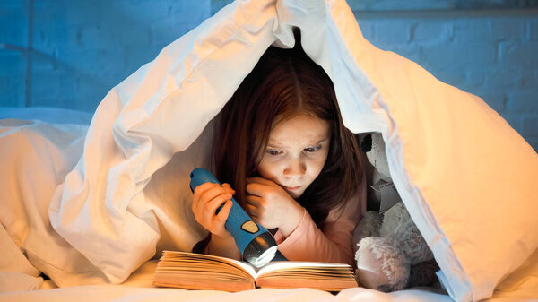 Focused kid with flashlight reading book under blanket on bed 