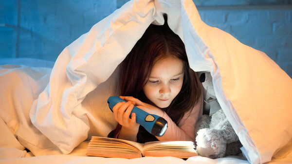 Preteen kid holding flashlight while reading book near soft toy on bed 