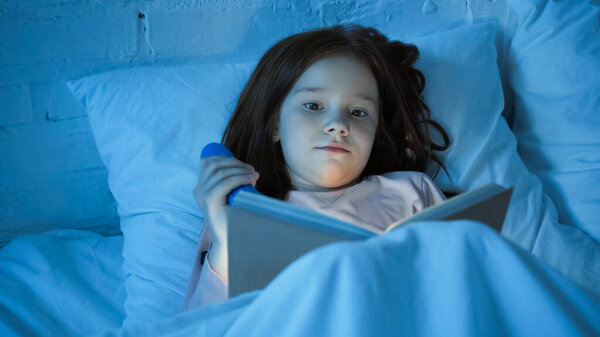 Child holding flashlight and reading book on blurred foreground on bed 