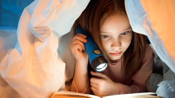 Kid holding flashlight and reading book under blanket in bedroom 