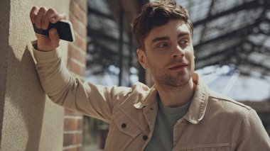 Bej ceketli yakışıklı adam akıllı telefon tutuyor ve alışveriş merkezinde başka tarafa bakıyor. 