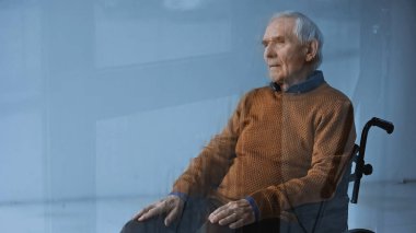 Tekerlekli sandalyedeki düşünceli yaşlı adam gri arka plana bakıyor.