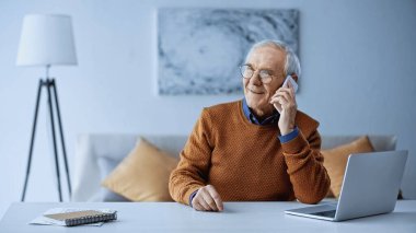 Neşeli yaşlı adam cep telefonuyla konuşuyor. Evde dizüstü bilgisayar var.