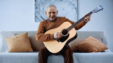 Oturma odasında gitarıyla kanepede oturan gülümseyen yaşlı adam.