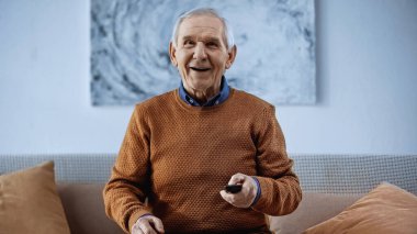 Kanepede elinde uzaktan kumandayla oturan ve evdeki kameraya bakan mutlu kıdemli bir adam.