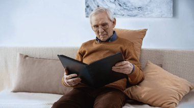elderly man sitting on sofa with family album at home clipart