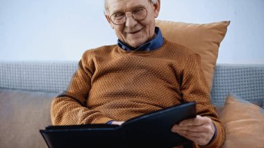 happy elderly man sitting on sofa with family album in living room clipart