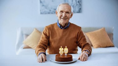 Neşeli yaşlı adam modern oturma odasında yanan mumlarla doğum günü pastasının önünde oturuyor.