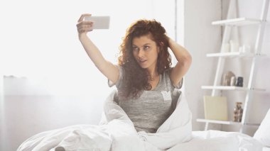 Kıvırcık genç kadın yatak odasında akıllı telefondan selfie çekiyor. 