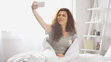 Mutlu genç bir kadın yatak odasında akıllı telefondan selfie çekiyor. 
