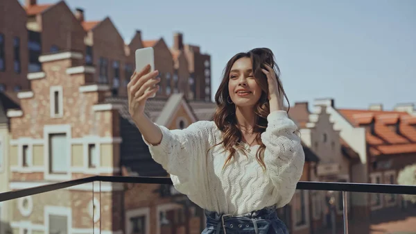 Mujer Joven Feliz Tomando Selfie Ajustando Cabello Cerca Edificios Borrosos — Foto de Stock