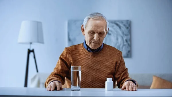 Anciano Sentado Cerca Mesa Con Frasco Medicina Vaso Agua Sala — Foto de Stock