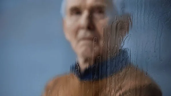 blurred portrait of elderly man looking at camera through rainy window on grey background