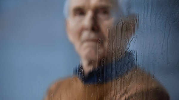blurred portrait of elderly man looking at camera through rainy window on grey background
