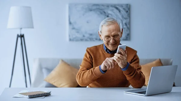 Uśmiechnięty Staruszek Siedzi Obok Laptopa Wysyła Wiadomości Smartfonie Domu — Zdjęcie stockowe