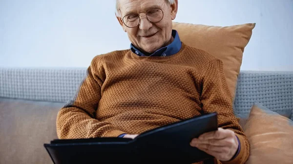 happy elderly man sitting on sofa with family album in living room