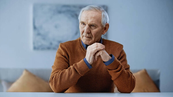serious elderly man with clenched hand near face sitting at table at home