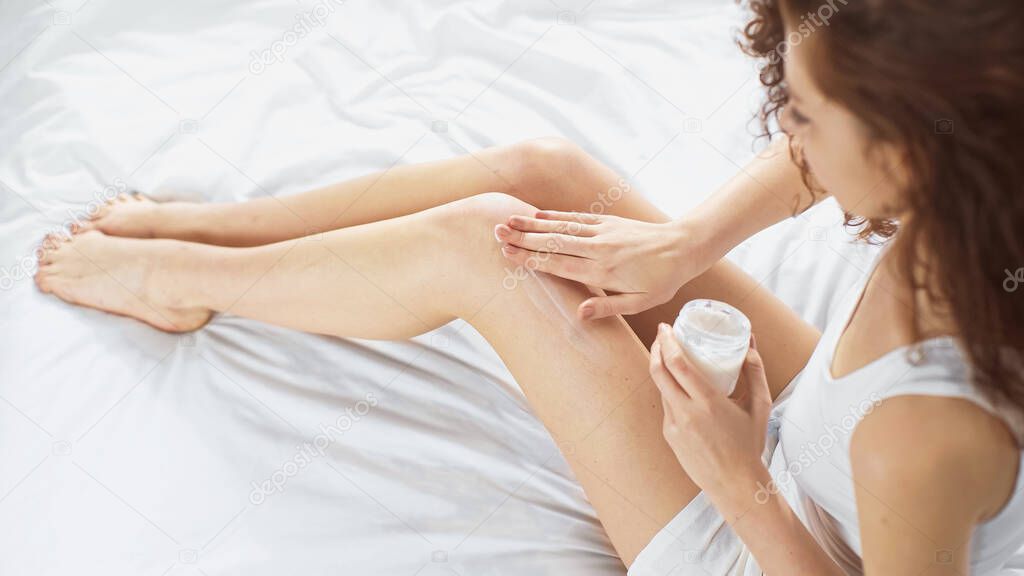 high angle view of curly young woman sitting on bed and applying body cream