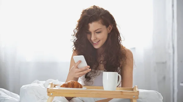 happy and curly woman holding cup of coffee and using smartphone near tray with croissant on bed