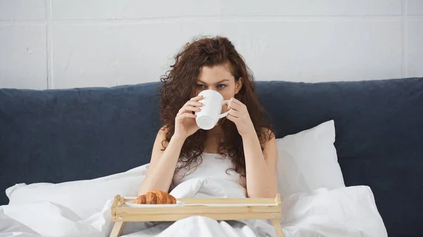 Giovane Donna Bere Caffè Vicino Vassoio Con Croissant Sul Letto — Foto Stock