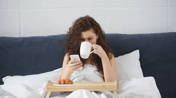 Jovem Mulher Encaracolado Beber Café Segurando Smartphone Perto Bandeja Com — Fotografia de Stock