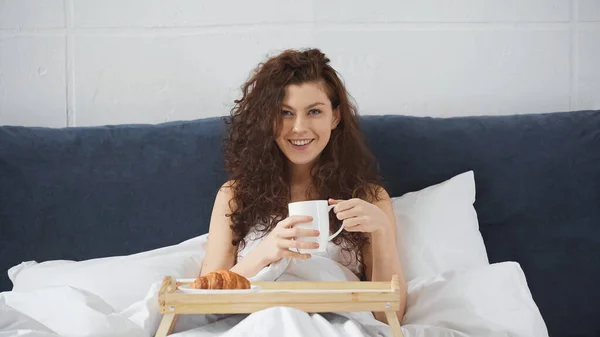 Lycklig Kvinna Håller Kopp Kaffe Nära Bricka Med Croissant Sängen — Stockfoto