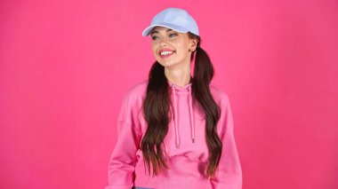 smiling young adult woman in cap and hoodie looking away isolated on pink