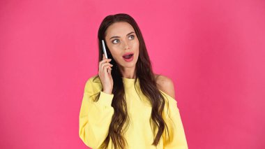 amazed young adult woman with open mouth speaking on cellphone isolated on pink