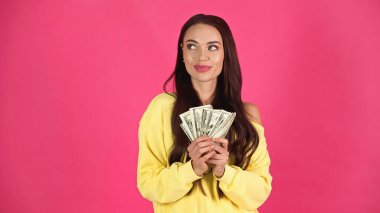 thoughtful young adult woman holding dollar banknotes in hands isolated on pink