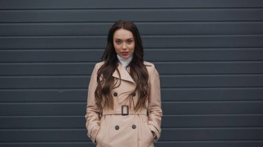 attractive young adult woman in beige trench coat and turtleneck looking at camera on textured grey background