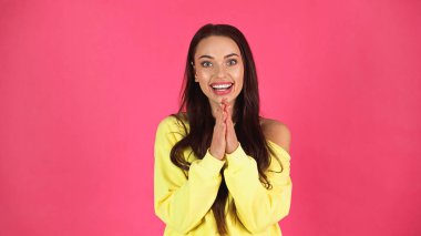 positive young adult woman in yellow blouse looking at camera with praying hands isolated on pink