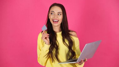 thoughtful young adult woman holding laptop and credit card isolated on pink