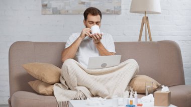 sick freelancer sitting on sofa with laptop and drinking tea near coffee table with pills in bottles 