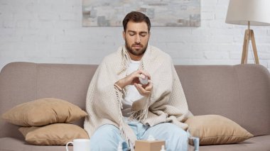 sick man sitting on sofa and holding bottle with pills near coffee table with drinks 