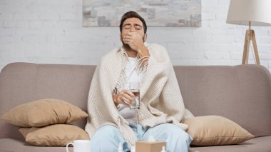 sick man sitting on sofa and taking pill near coffee table with cup and bottles 