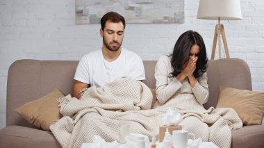 sick woman sneezing near man and bottles with pills on coffee table 
