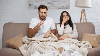 sick man holding glass of water near diseased woman sneezing in living room 