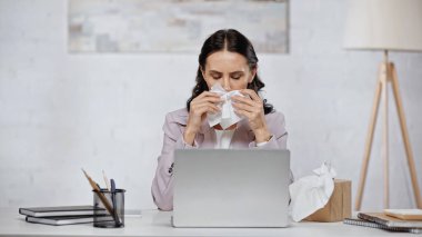 brunette and allergic businesswoman sneezing in tissue near laptop on desk