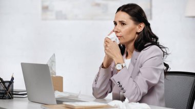 allergic businesswoman sneezing in napkin near laptop in office