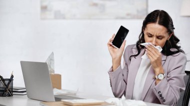 allergic businesswoman sneezing in tissue near laptop while holding smartphone