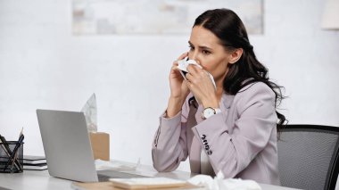 brunette businesswoman with allergy sneezing in napkin while talking on cellphone near laptop on desk