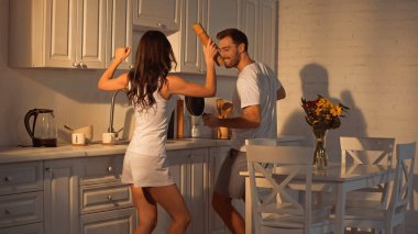cheerful man with frying pan dancing near girlfriend with paper mill   clipart