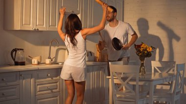 happy man with frying pan and wooden spoon dancing near girlfriend with paper mill   clipart
