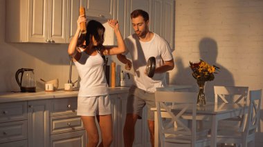happy man with frying pan dancing near brunette girlfriend with paper mill  