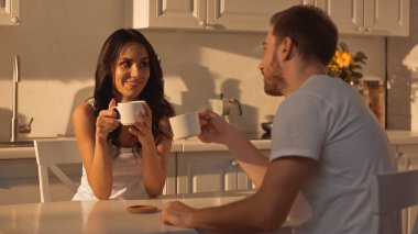 smiling young woman holding cup of coffee and talking with blurred boyfriend 