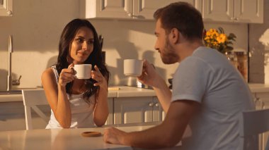 young woman holding cup of coffee and smiling near blurred boyfriend 