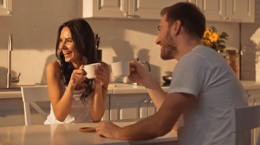 happy young woman holding cup and smiling near blurred boyfriend 