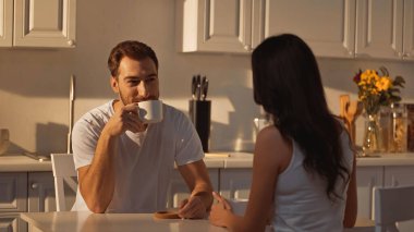 happy man drinking coffee and looking at brunette girlfriend in kitchen 