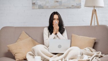 ill woman sneezing in tissue while looking at laptop 