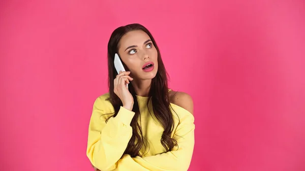 thoughtful young adult woman with open mouth speaking on cellphone isolated on pink